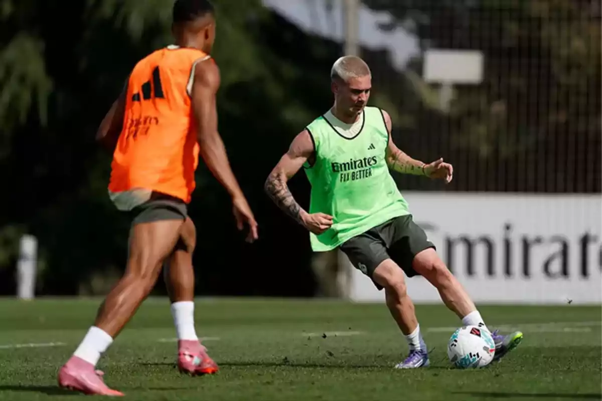 Dos futbolistas entrenan en una cancha de césped uno con peto verde controla el balón mientras el otro con peto naranja se acerca para defender