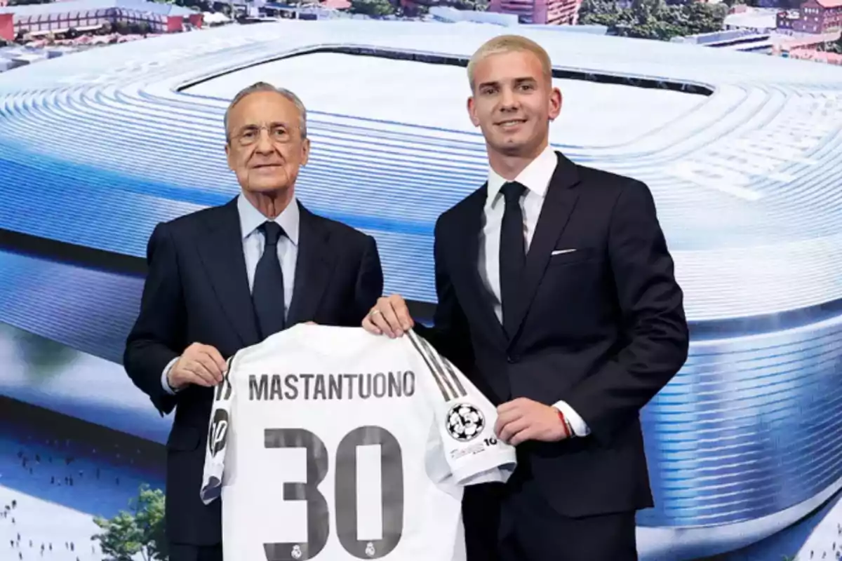 Two men in suits pose smiling, holding a soccer jersey with the name Mastantuono and the number 30 in front of an image of Santiago Bernabéu stadium. Two men in suits pose smiling, holding a soccer jersey with the name Mastantuono and the number 30 in front of an image of Santiago Bernabéu stadium.