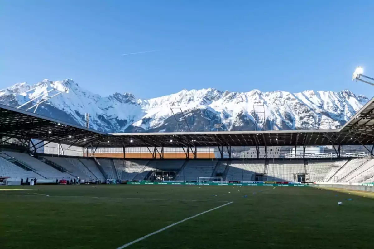 Estadio de fútbol vacío con gradas y techo, al fondo montañas nevadas bajo un cielo despejado Estadio de fútbol vacío con gradas y techo, al fondo montañas nevadas bajo un cielo despejado
