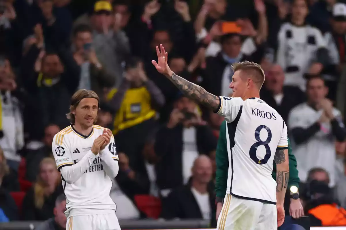 Dos futbolistas del Real Madrid en el campo, uno aplaudiendo y el otro levantando la mano mientras la afición los observa desde las gradas