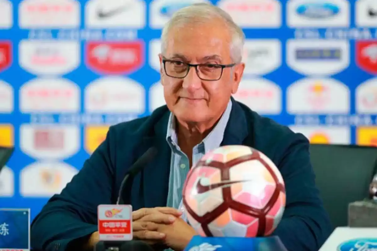 Hombre de cabello canoso y gafas sentado en una conferencia de prensa de fútbol con un balón y micrófonos frente a él Hombre de cabello canoso y gafas sentado en una conferencia de prensa de fútbol con un balón y micrófonos frente a él
