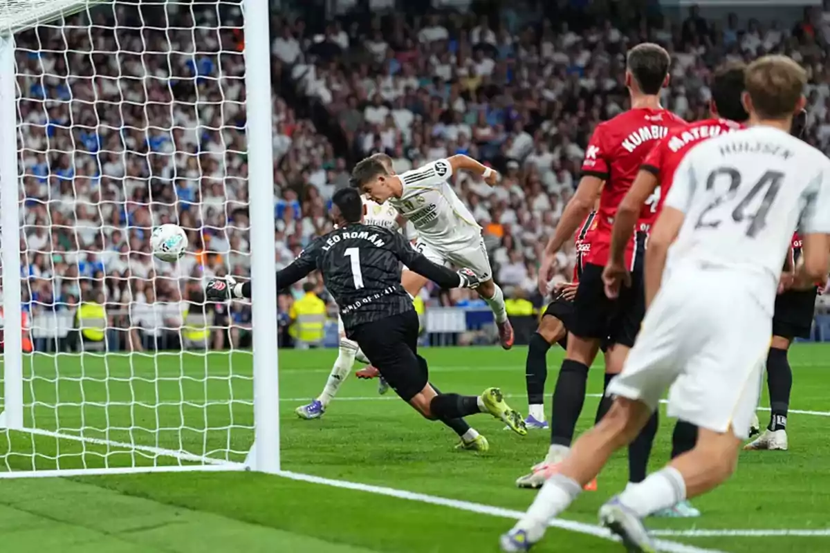 Un jugador de fútbol cabecea el balón hacia la portería mientras el portero y varios defensores intentan evitar el gol en un estadio lleno de espectadores. Un jugador de fútbol cabecea el balón hacia la portería mientras el portero y varios defensores intentan evitar el gol en un estadio lleno de espectadores.
