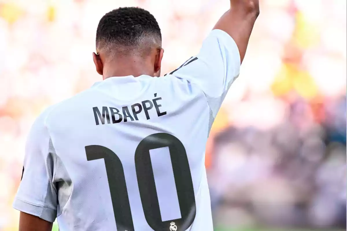 Jugador de fútbol con la camiseta blanca número 10 y el nombre Mbappé levantando el brazo en un estadio lleno