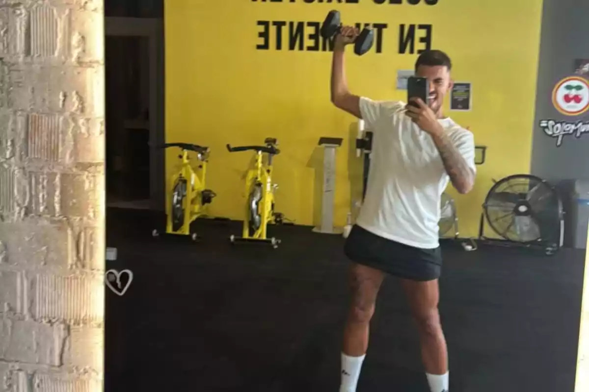 Hombre tomándose una selfie en el gimnasio mientras sostiene una mancuerna con una mano y lleva puesta una camiseta blanca y una falda negra