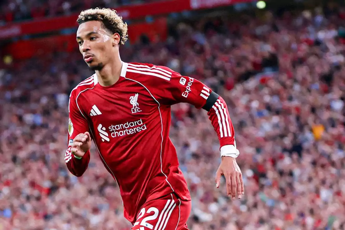 Futbolista con uniforme rojo del Liverpool corriendo en el campo durante un partido con el estadio lleno de aficionados al fondo Futbolista con uniforme rojo del Liverpool corriendo en el campo durante un partido con el estadio lleno de aficionados al fondo
