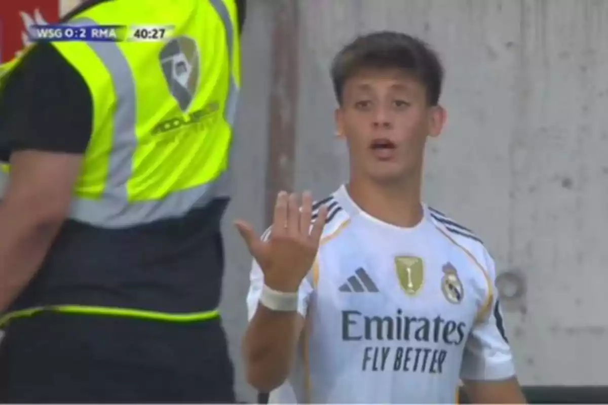 Real Madrid player with a surprised or protesting expression during a match, wearing the team’s white uniform and making a hand gesture while a security guard stands with his back next to him.
