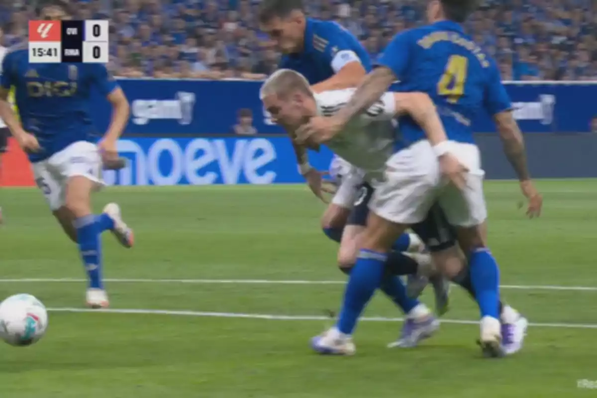 Jugadores de fútbol disputan el balón en el área durante un partido mientras uno de ellos es derribado por un rival con la camiseta azul Jugadores de fútbol disputan el balón en el área durante un partido mientras uno de ellos es derribado por un rival con la camiseta azul