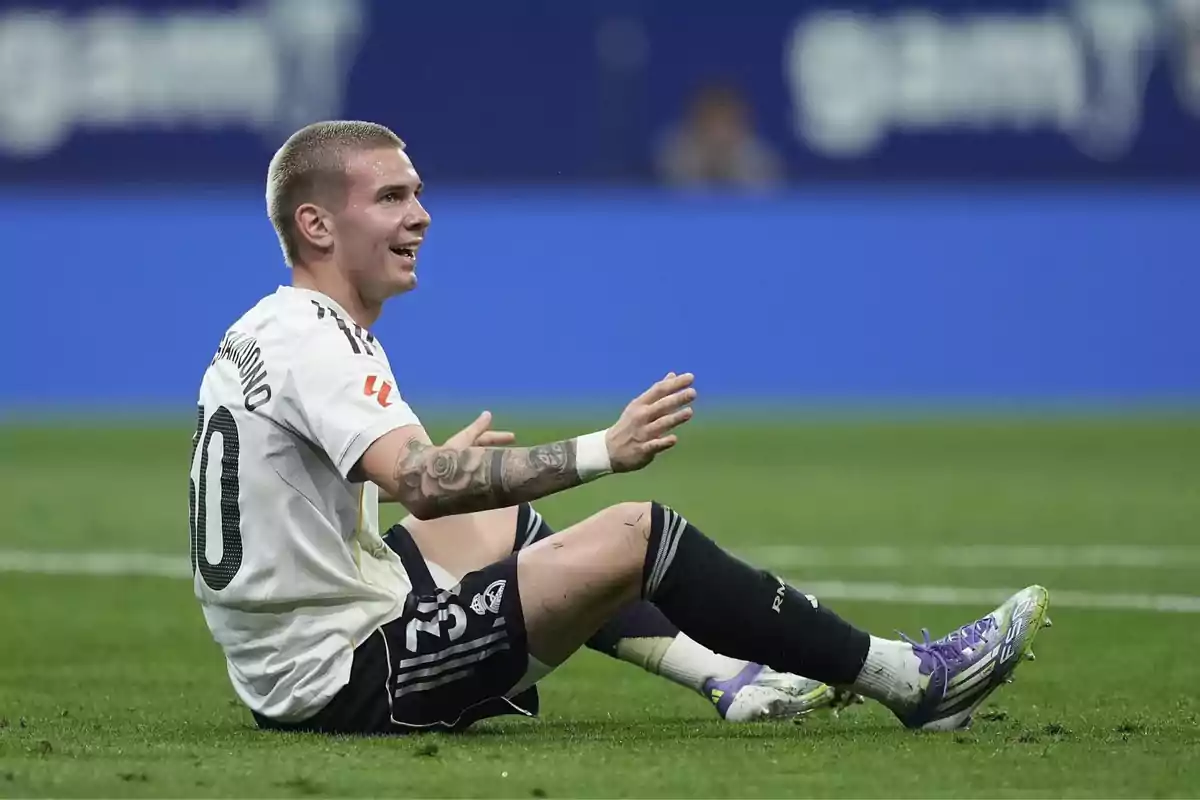 Futbolista sentado en el césped durante un partido con expresión sonriente y uniforme blanco Futbolista sentado en el césped durante un partido con expresión sonriente y uniforme blanco
