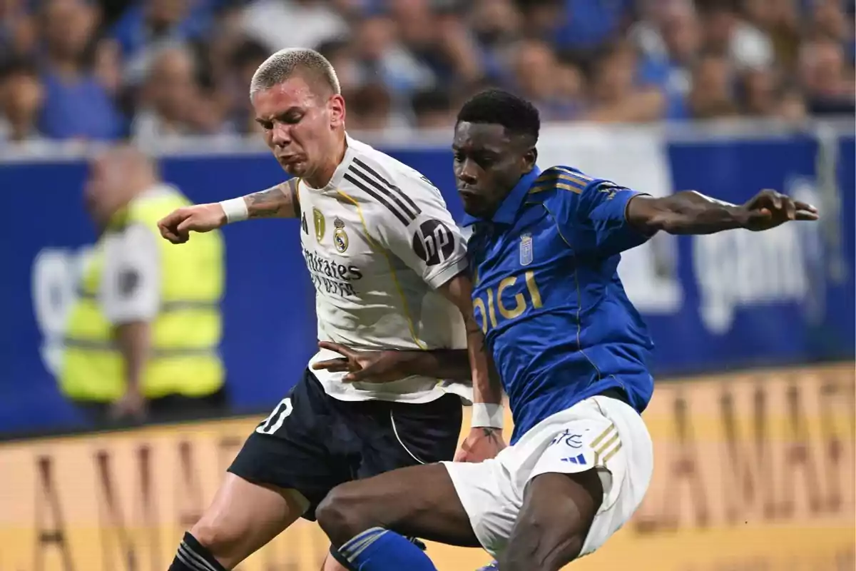 Dos futbolistas disputan el balón durante un partido uno con uniforme blanco y el otro con uniforme azul mientras el público observa desde las gradas