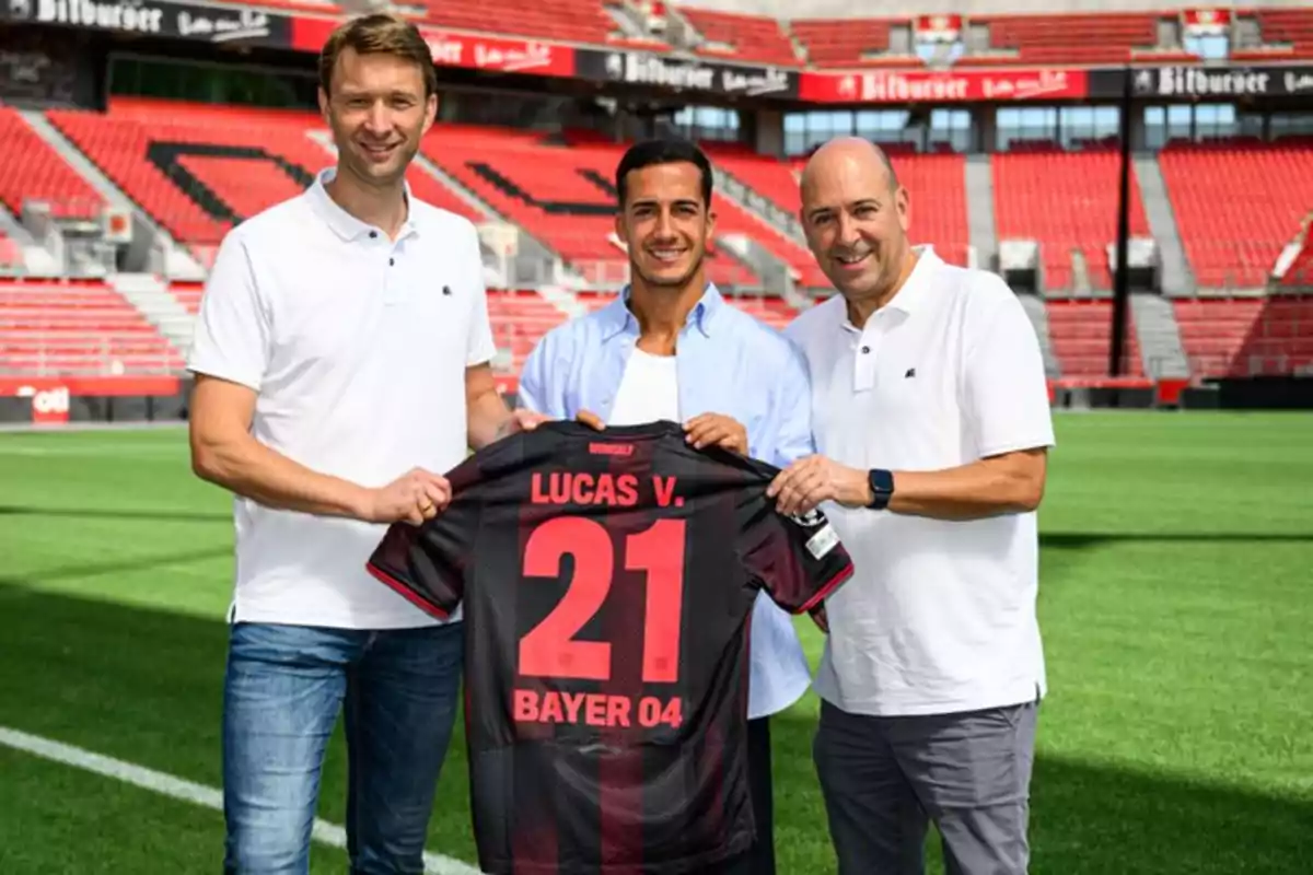 Three men pose in a soccer stadium holding a Bayer 04 jersey with the name Lucas V. and the number 21. Three men pose in a soccer stadium holding a Bayer 04 jersey with the name Lucas V. and the number 21.