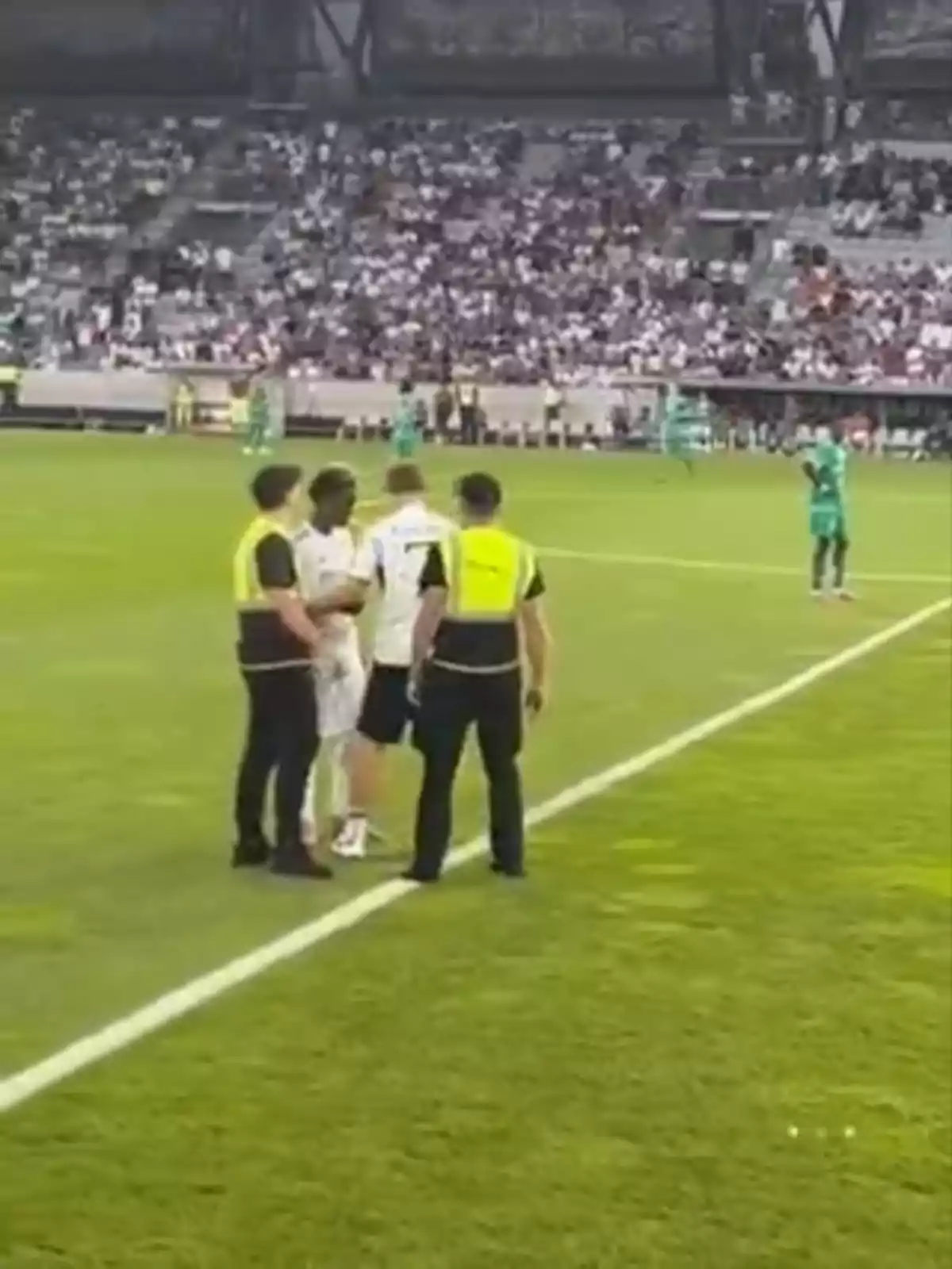 Un grupo de guardias de seguridad detiene a una persona vestida de blanco en el borde de una cancha de fútbol mientras el público observa desde las gradas. Un grupo de guardias de seguridad detiene a una persona vestida de blanco en el borde de una cancha de fútbol mientras el público observa desde las gradas.