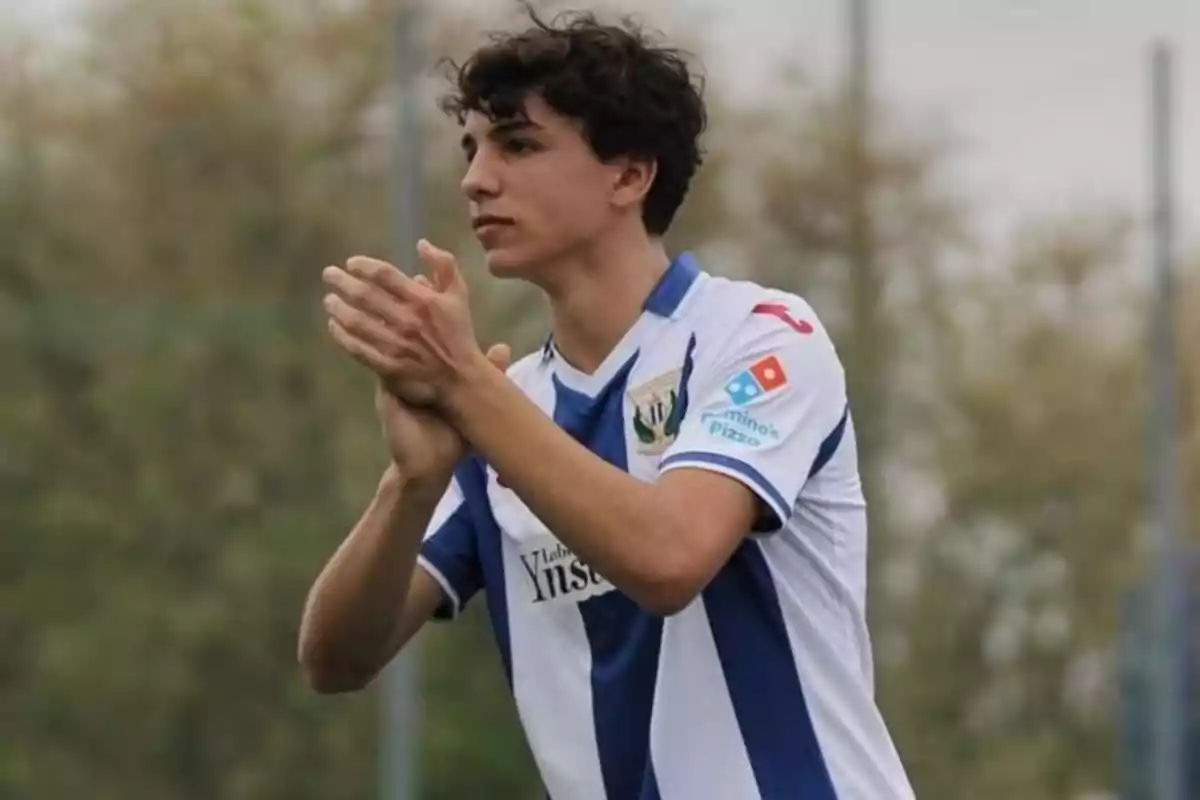 Joven futbolista con uniforme blanco y azul aplaudiendo en un campo al aire libre Joven futbolista con uniforme blanco y azul aplaudiendo en un campo al aire libre