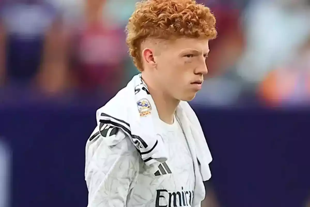 Young player with curly reddish hair, wearing a white jersey and a towel over his shoulders in a stadium Young player with curly reddish hair, wearing a white jersey and a towel over his shoulders in a stadium