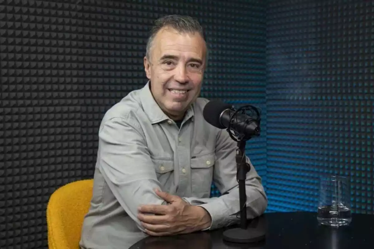 Hombre de mediana edad sonriente sentado frente a un micrófono en un estudio de grabación con paredes insonorizadas y una silla amarilla Hombre de mediana edad sonriente sentado frente a un micrófono en un estudio de grabación con paredes insonorizadas y una silla amarilla