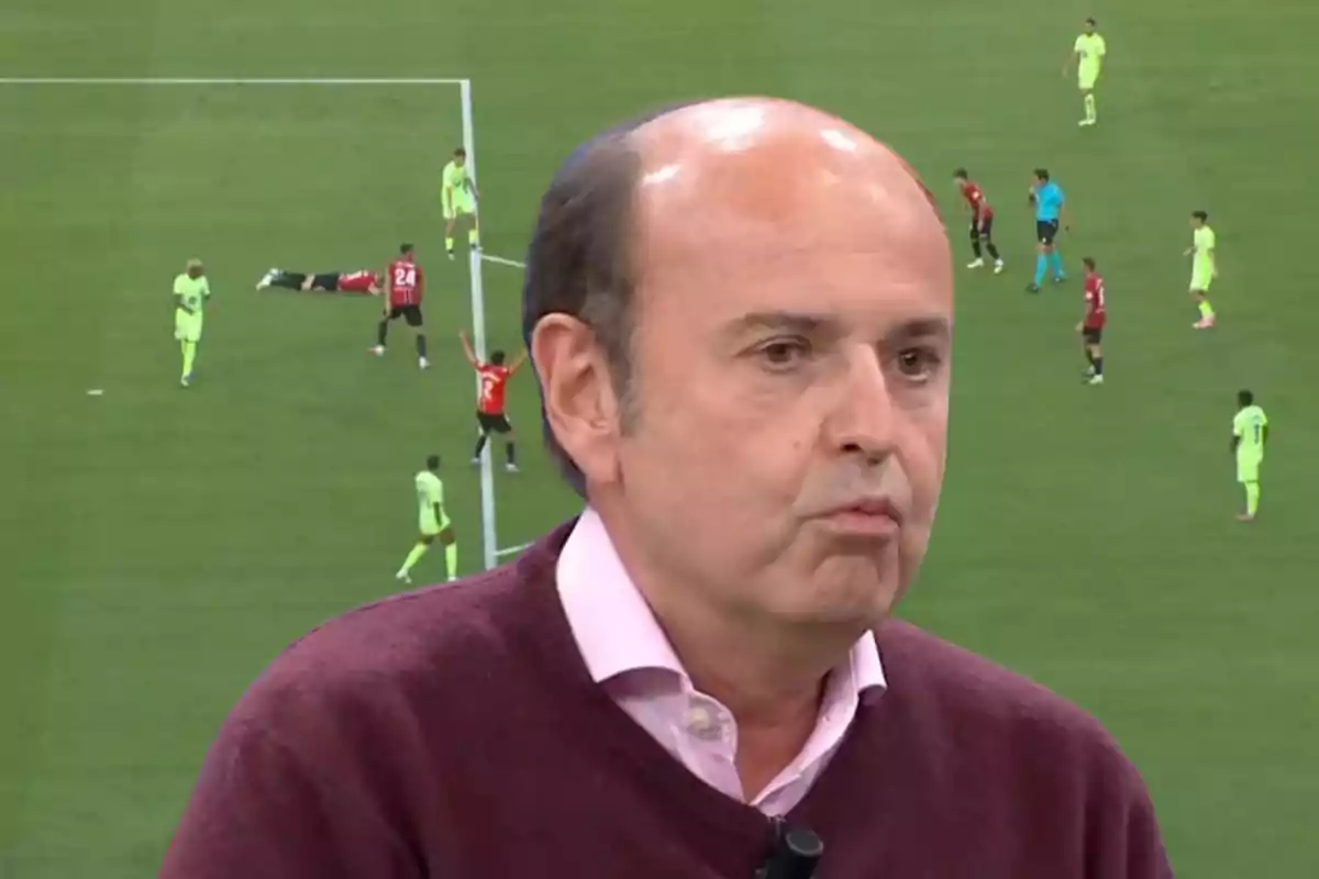 Un hombre calvo con suéter burdeos y camisa rosa aparece en primer plano con una expresión seria mientras de fondo se observa una escena de un partido de fútbol con jugadores en el campo
