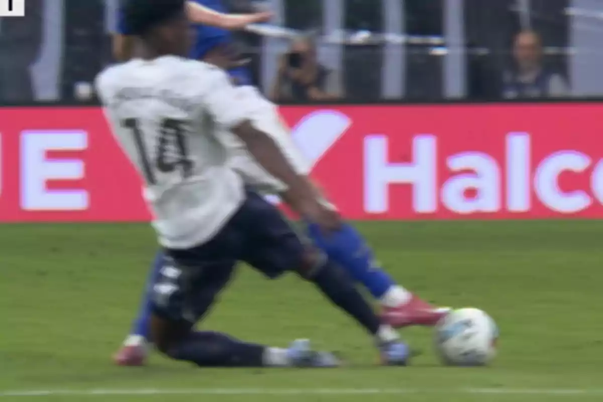 Jugador con camiseta blanca realiza una barrida para intentar quitarle el balón a un oponente vestido de azul durante un partido de fútbol Jugador con camiseta blanca realiza una barrida para intentar quitarle el balón a un oponente vestido de azul durante un partido de fútbol
