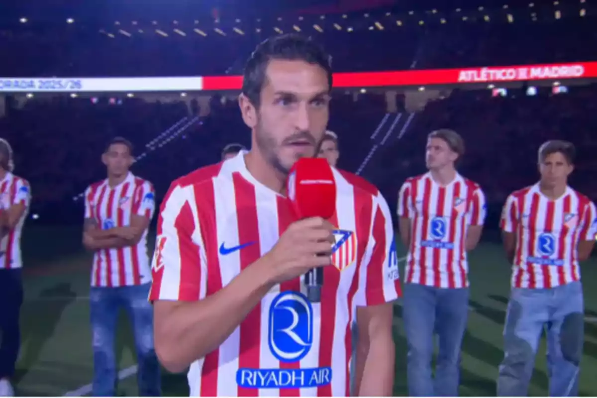 Un futbolista del Atlético de Madrid sostiene un micrófono rojo mientras da un discurso en un estadio acompañado de varios compañeros con el mismo uniforme. Un futbolista del Atlético de Madrid sostiene un micrófono rojo mientras da un discurso en un estadio acompañado de varios compañeros con el mismo uniforme.