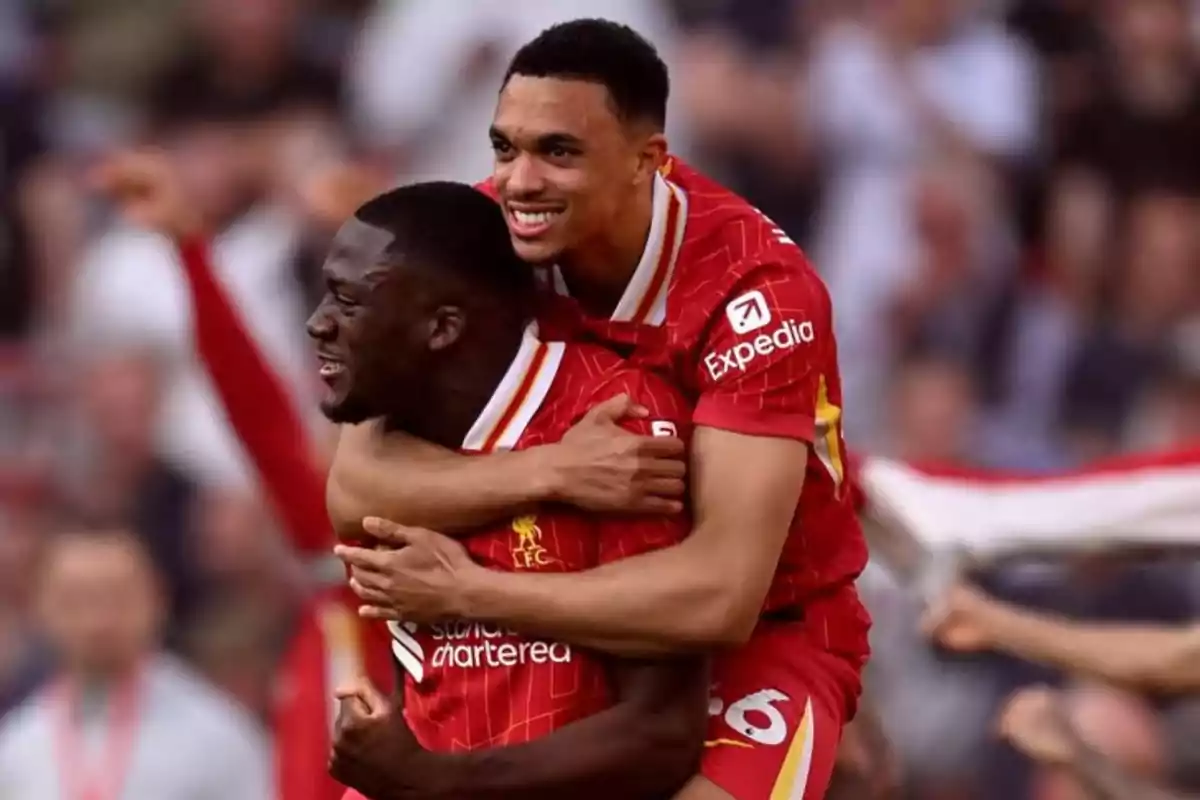 Two Liverpool footballers celebrate a goal by hugging on the field with expressions of joy and excitement while the crowd watches them in the background. Two Liverpool footballers celebrate a goal by hugging on the field with expressions of joy and excitement while the crowd watches them in the background.