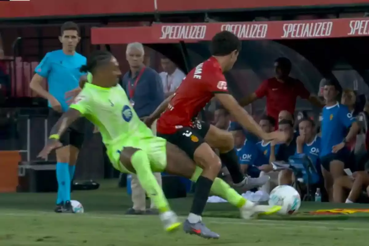 Un jugador con uniforme fosforescente realiza una barrida sobre un jugador con uniforme rojo durante un partido de fútbol mientras el árbitro y el cuerpo técnico observan la jugada desde la banda Un jugador con uniforme fosforescente realiza una barrida sobre un jugador con uniforme rojo durante un partido de fútbol mientras el árbitro y el cuerpo técnico observan la jugada desde la banda