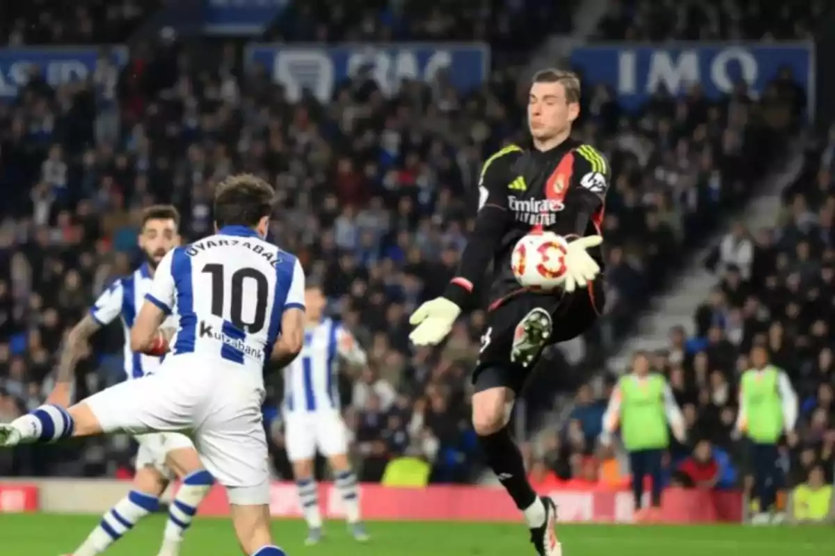 Portero vestido de negro atajando un balón mientras un jugador con el número 10 de un equipo rival intenta rematar durante un partido de fútbol con público en las gradas Portero vestido de negro atajando un balón mientras un jugador con el número 10 de un equipo rival intenta rematar durante un partido de fútbol con público en las gradas