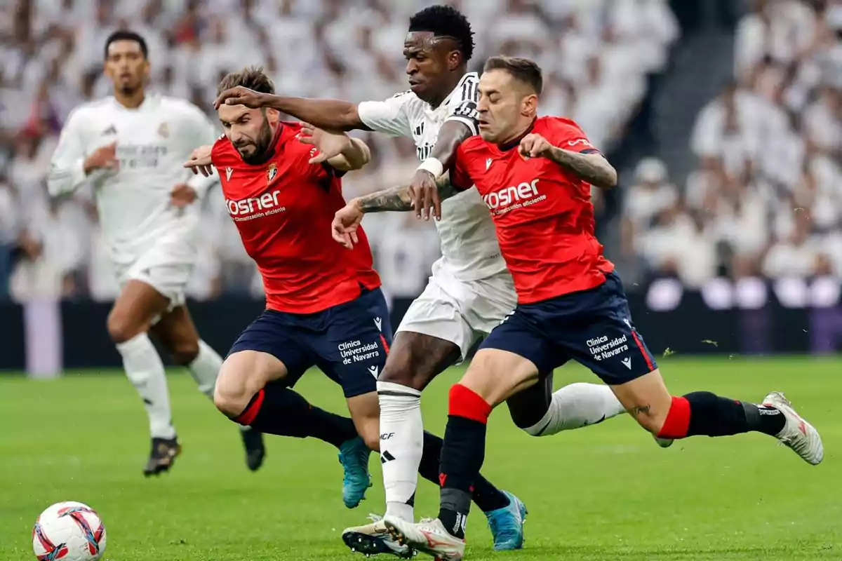 Three players compete for the ball during a match; two players in red uniforms and one in a white uniform run intensely on the field. Three players compete for the ball during a match; two players in red uniforms and one in a white uniform run intensely on the field.