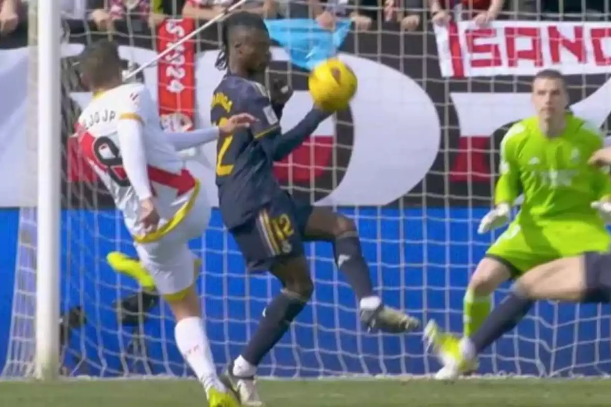 Un jugador de fútbol detiene el balón con la mano dentro del área mientras un rival intenta rematar y el portero observa la jugada. Un jugador de fútbol detiene el balón con la mano dentro del área mientras un rival intenta rematar y el portero observa la jugada.