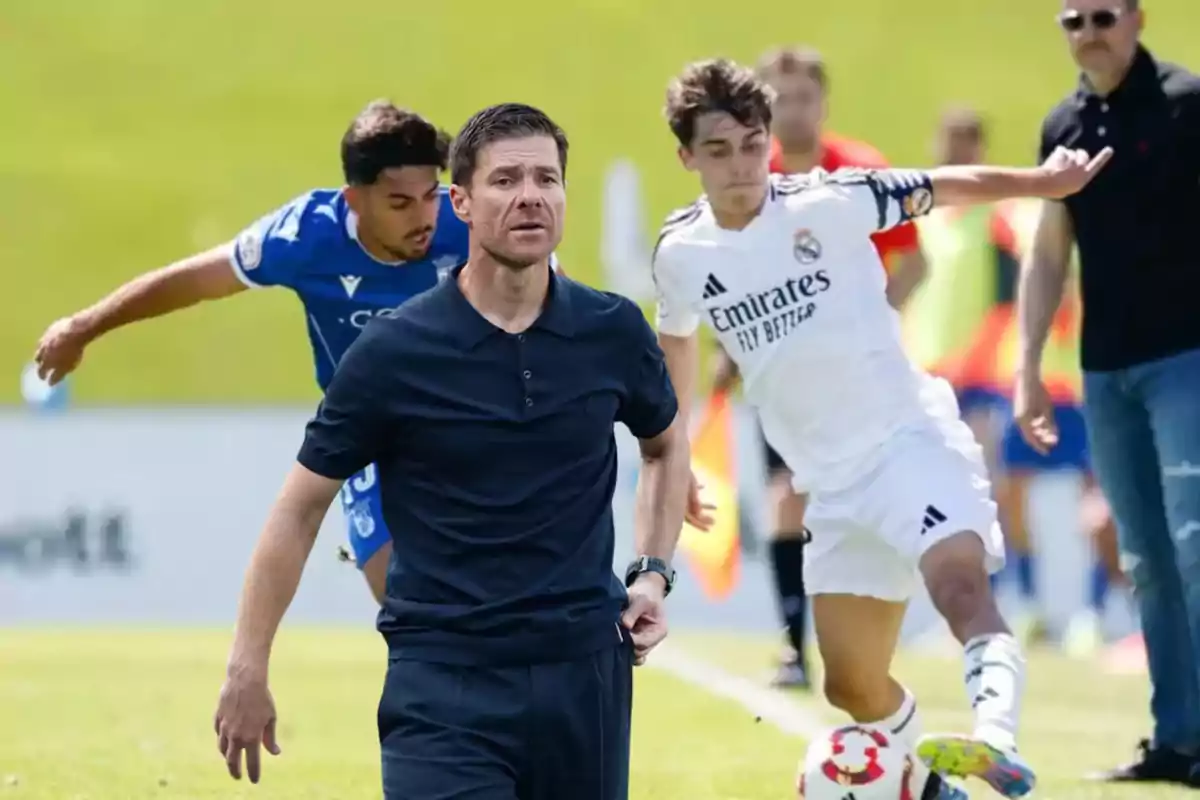 Un entrenador de fútbol con camisa azul oscuro aparece en primer plano mientras al fondo dos jugadores disputan el balón en un partido, uno con uniforme blanco y otro con uniforme azul.