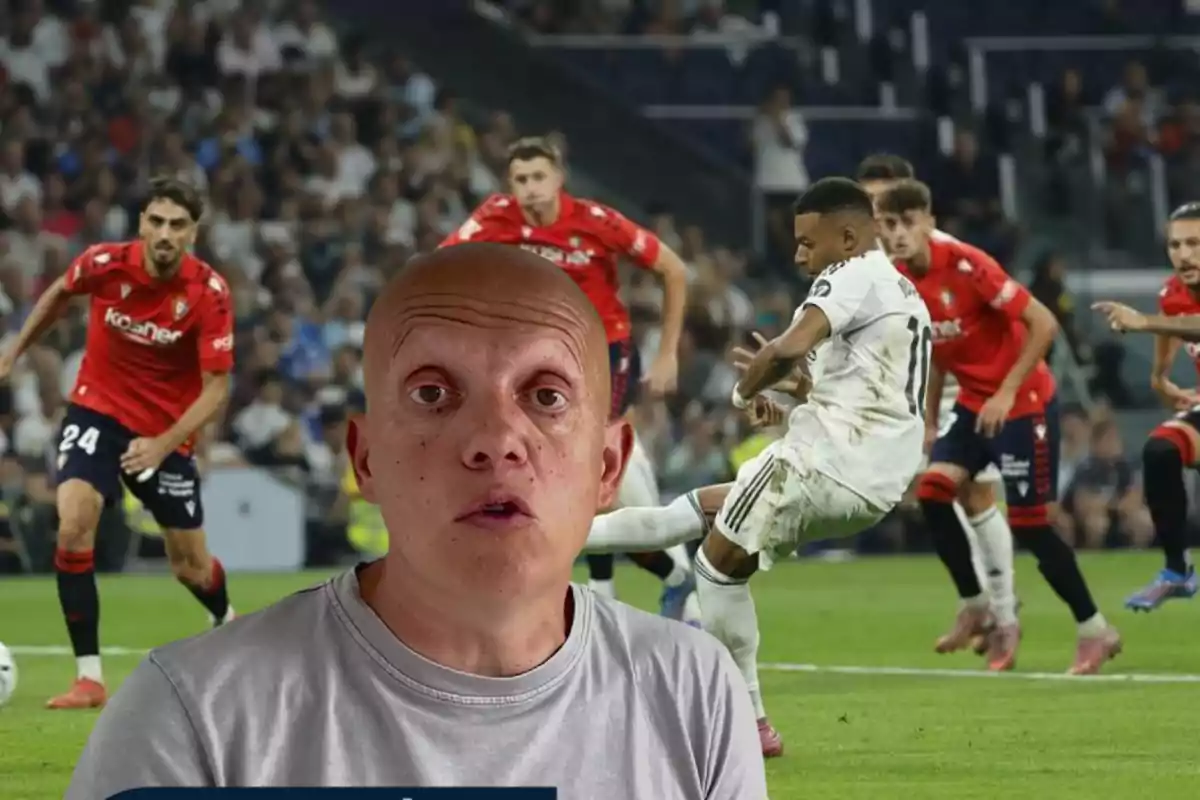 A bald man in the foreground with a serious expression, and in the background a soccer match where a player in white kicks the ball while several players in red watch him.