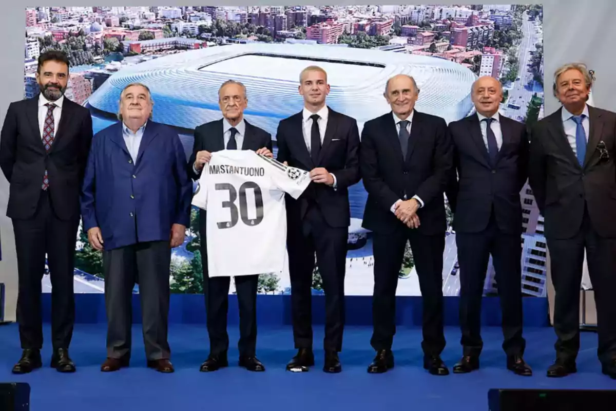 A group of men in suits pose for a photo while one of them holds a white soccer jersey with the name Mastantuono and the number 30; in the background, an image of a modern stadium can be seen. A group of men in suits pose for a photo while one of them holds a white soccer jersey with the name Mastantuono and the number 30; in the background, an image of a modern stadium can be seen.