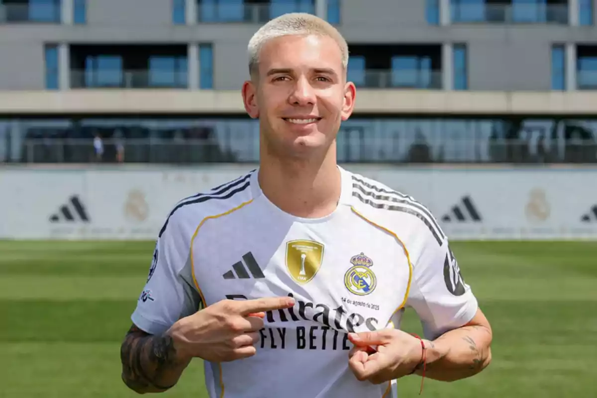 Jugador de fútbol con cabello corto y rubio sonríe mientras señala el escudo del Real Madrid en su camiseta blanca durante el día en un campo de entrenamiento Jugador de fútbol con cabello corto y rubio sonríe mientras señala el escudo del Real Madrid en su camiseta blanca durante el día en un campo de entrenamiento