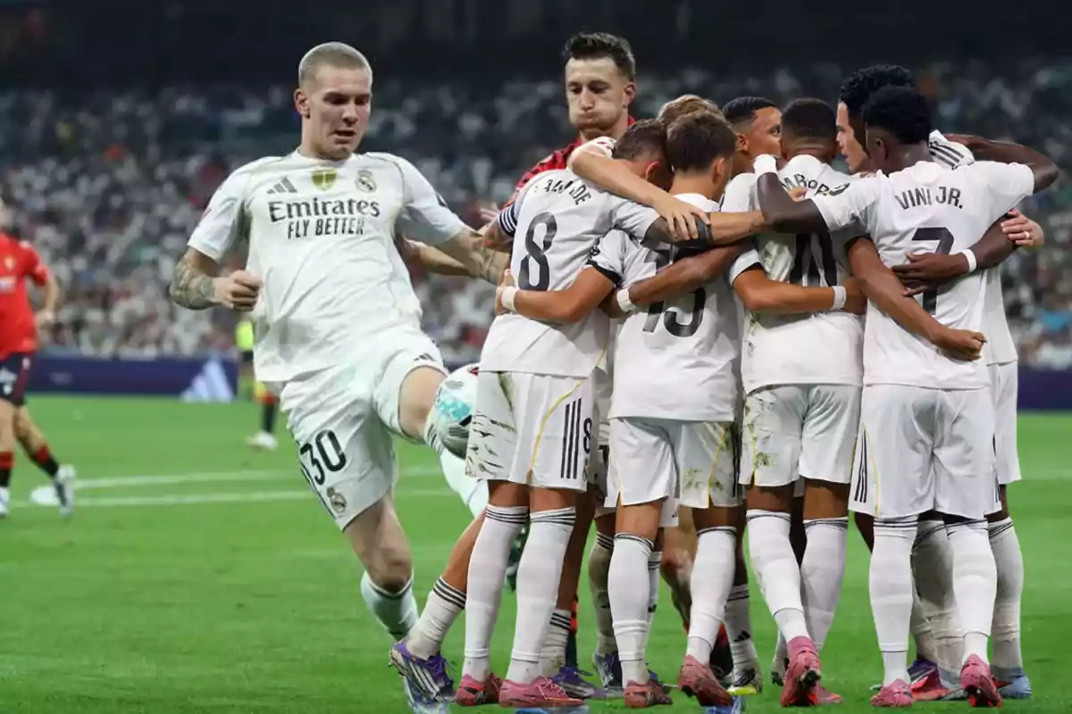 Un jugador de fútbol del Real Madrid controla el balón mientras un grupo de sus compañeros se abrazan celebrando en el campo
