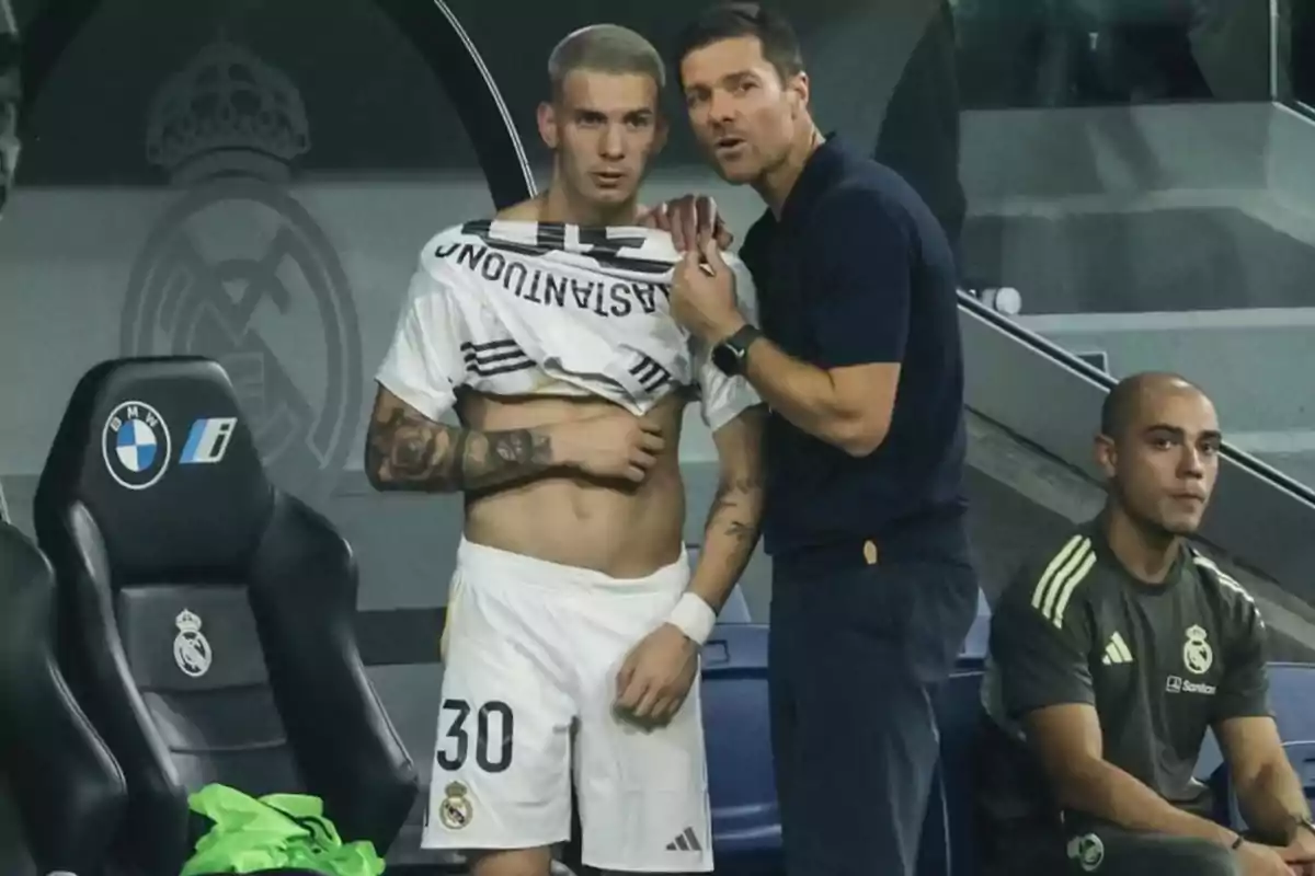 Soccer coach gives instructions to a player who is lifting his jersey while another team member watches from the bench.