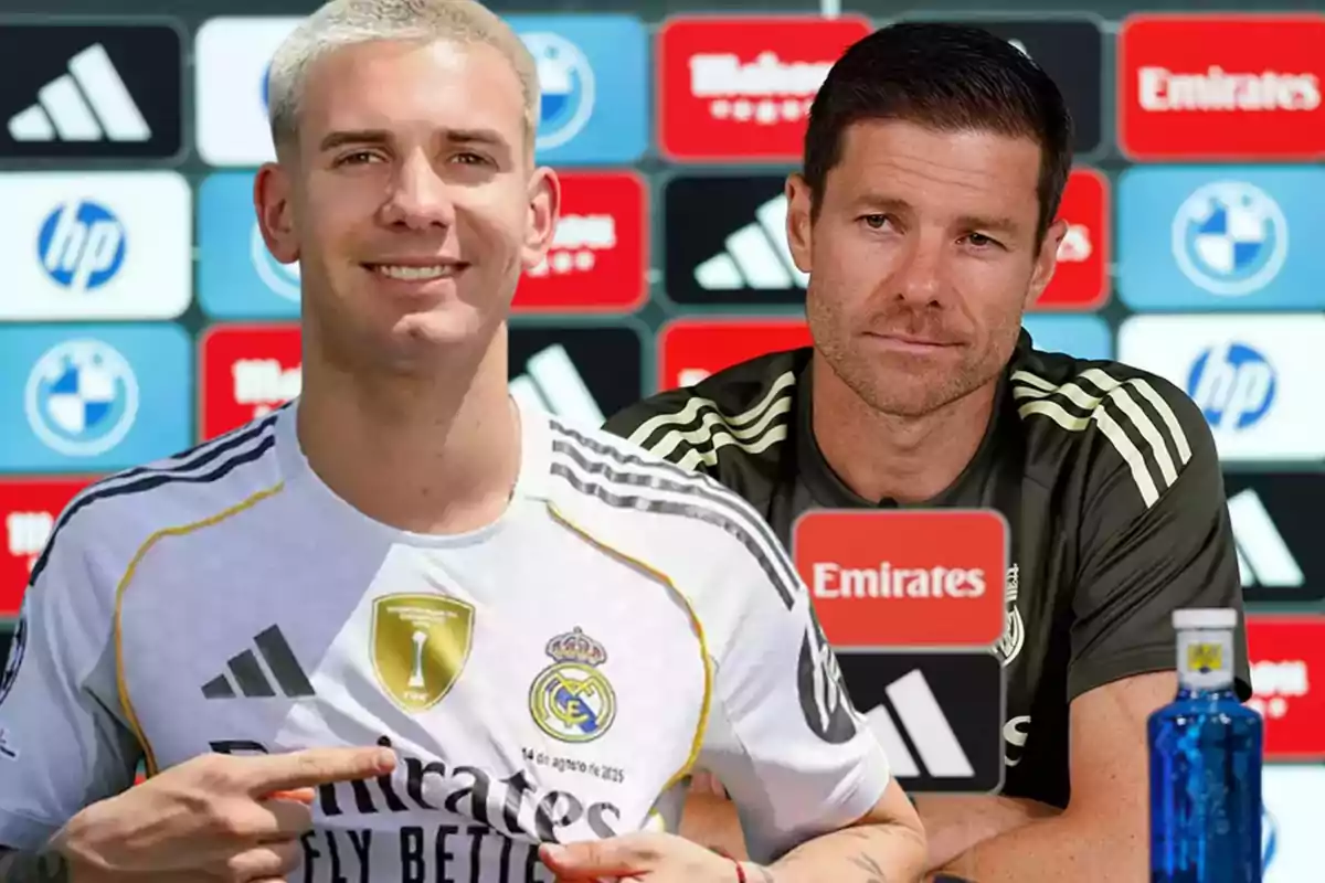 A player wearing a Real Madrid jersey smiles and points to the team’s badge while a coach sits in the background at a press conference.