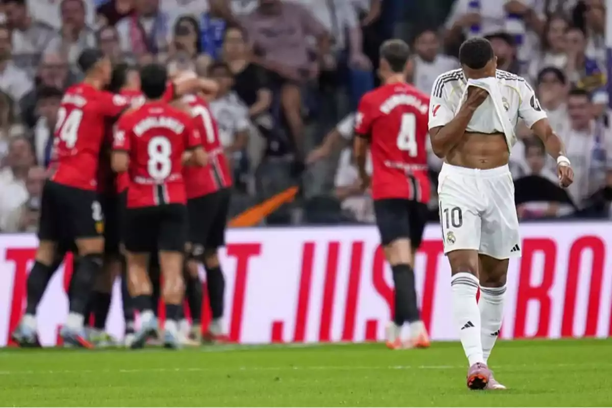 A Real Madrid player covers his face with his jersey while players from the opposing team celebrate a goal in the background. A Real Madrid player covers his face with his jersey while players from the opposing team celebrate a goal in the background.