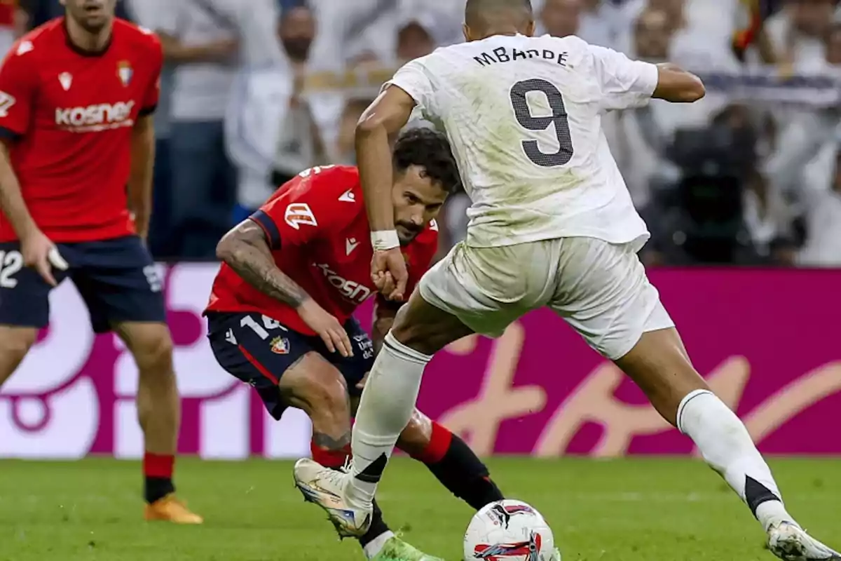 Jugador con uniforme blanco y el número 9 enfrentando a un defensor con uniforme rojo durante un partido de fútbol
