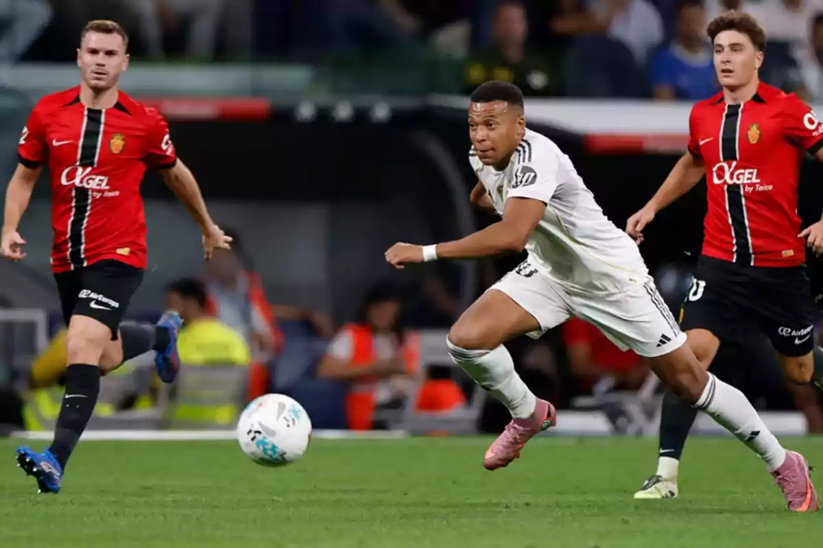 Tres futbolistas disputan el balón durante un partido, dos con uniforme rojo y negro y uno con uniforme blanco, en un estadio lleno de espectadores. Tres futbolistas disputan el balón durante un partido, dos con uniforme rojo y negro y uno con uniforme blanco, en un estadio lleno de espectadores.