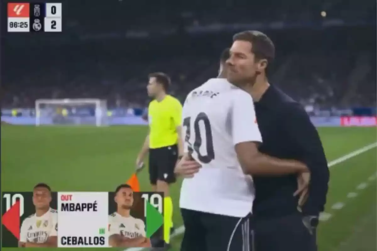 Un jugador de fútbol con el número 10 abraza a un entrenador en la banda mientras se realiza un cambio y el marcador muestra que el equipo visitante gana 2 a 0.