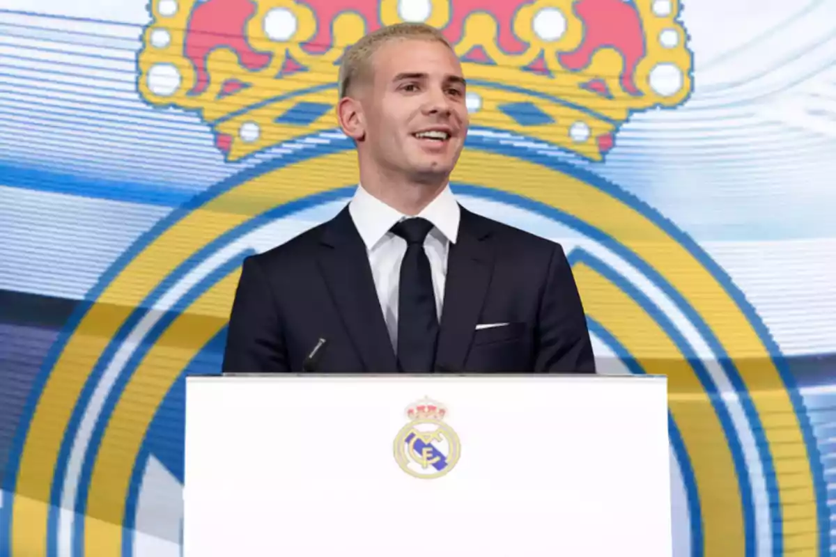 Un hombre con cabello corto y rubio vestido con traje oscuro y corbata negra sonríe mientras habla en un podio con el escudo del Real Madrid al fondo.