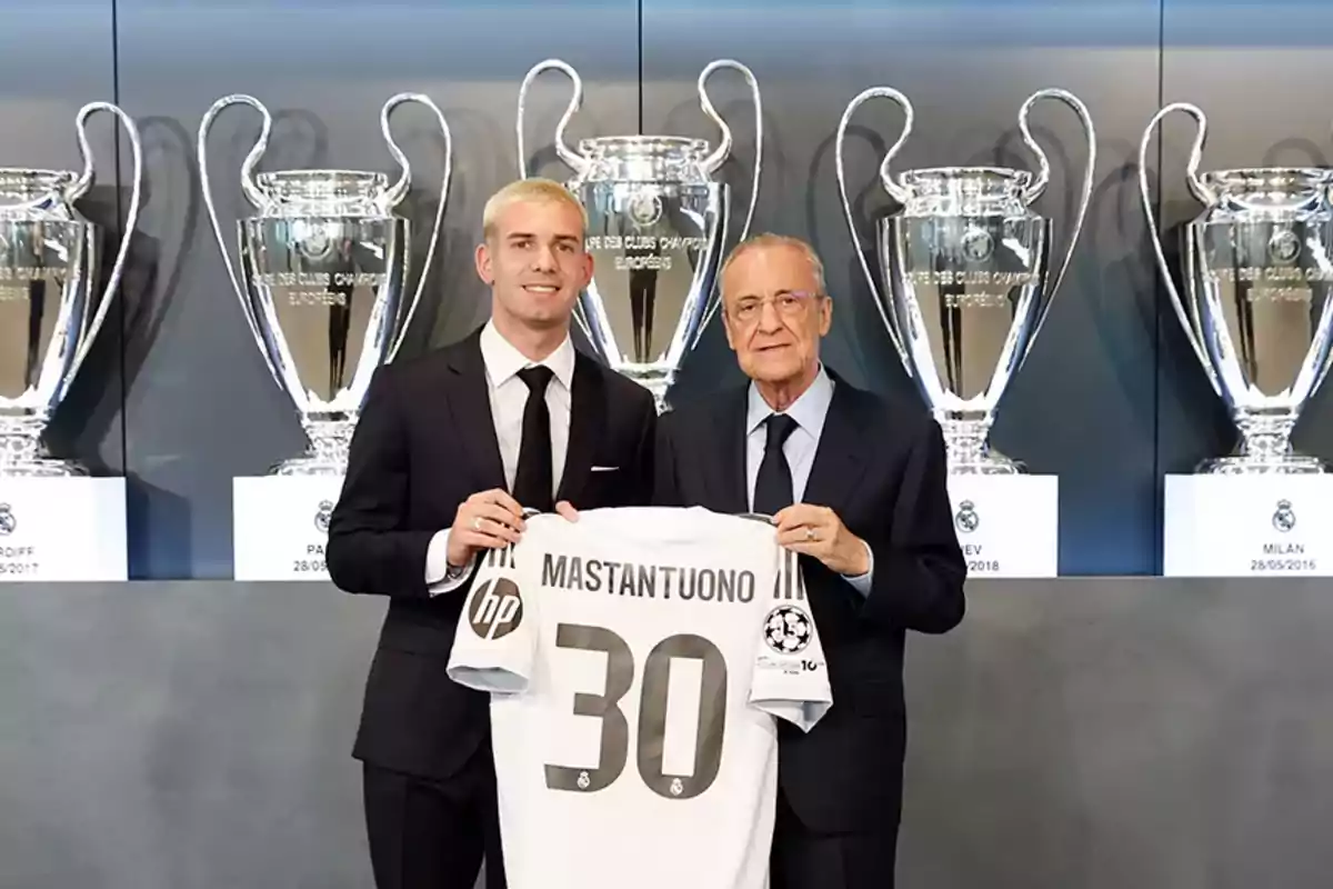 Dos hombres con traje sostienen una camiseta de fútbol con el nombre Mastantuono y el número 30 frente a varios trofeos grandes de la Champions League. Dos hombres con traje sostienen una camiseta de fútbol con el nombre Mastantuono y el número 30 frente a varios trofeos grandes de la Champions League.