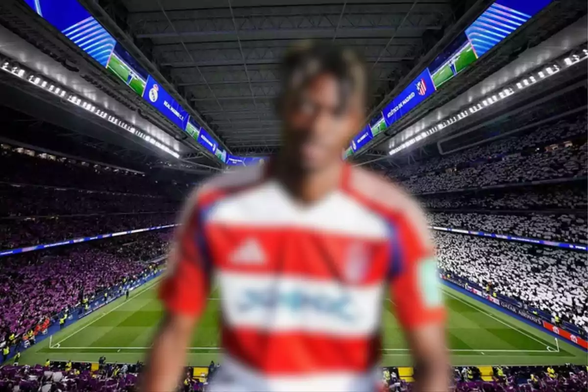 A player with a blurred face wears a red and white uniform while standing in front of a stadium full of fans.