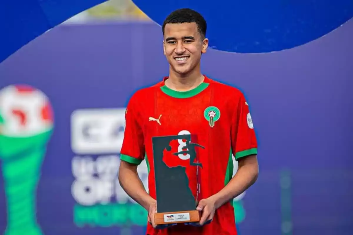 Jugador de fútbol con uniforme rojo de Marruecos sonríe mientras sostiene un trofeo rectangular con la silueta del continente africano sobre un fondo azul Jugador de fútbol con uniforme rojo de Marruecos sonríe mientras sostiene un trofeo rectangular con la silueta del continente africano sobre un fondo azul