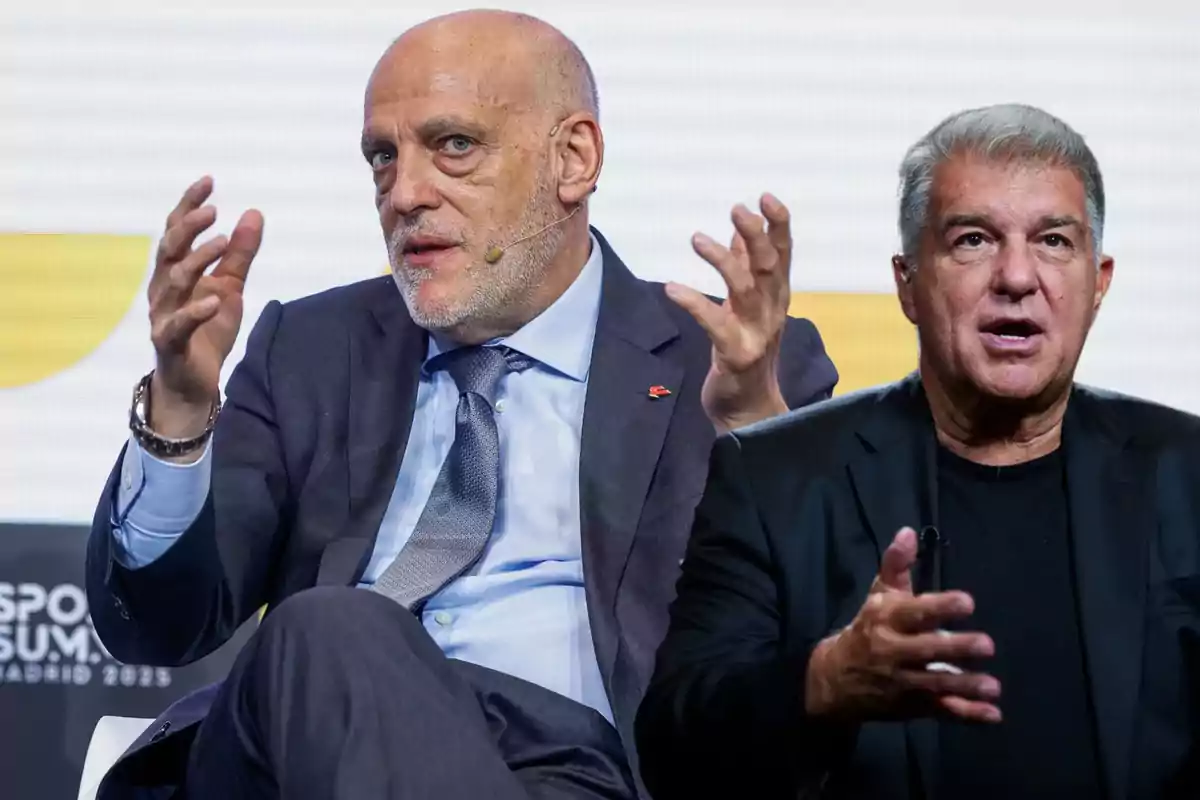 Two middle-aged men are talking at an event, both gesturing with their hands while seated and wearing formal clothing.