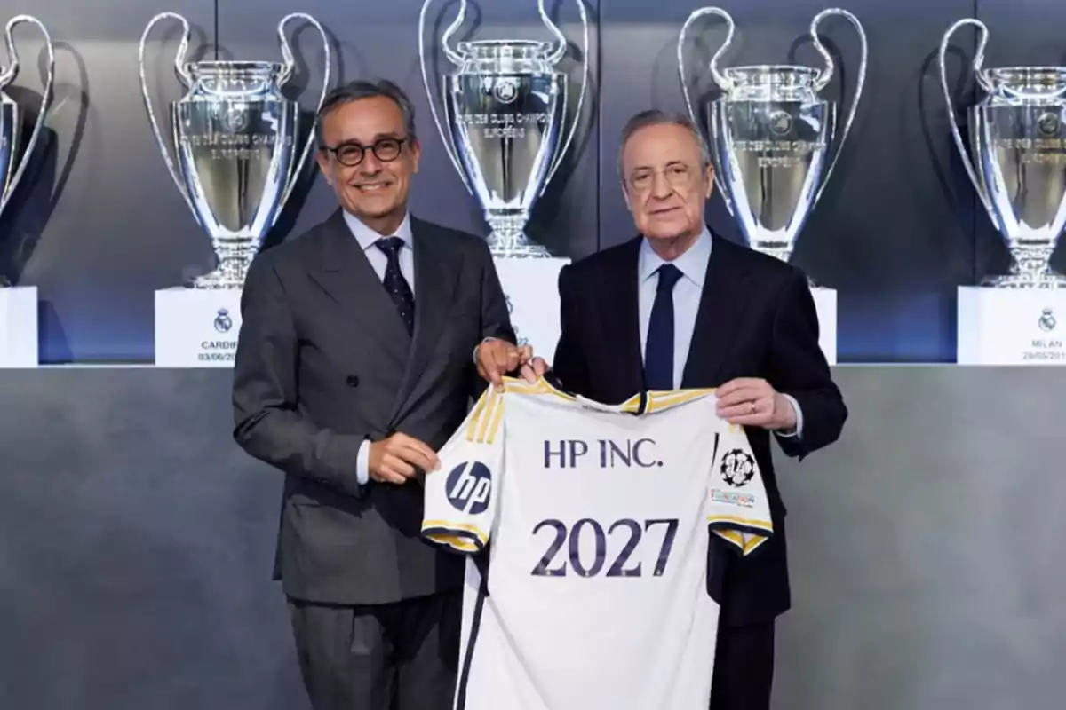 Two men in suits pose smiling while holding a white soccer jersey with the text HP INC and 2027; in the background, several large trophy cups can be seen. Two men in suits pose smiling while holding a white soccer jersey with the text HP INC and 2027; in the background, several large trophy cups can be seen.