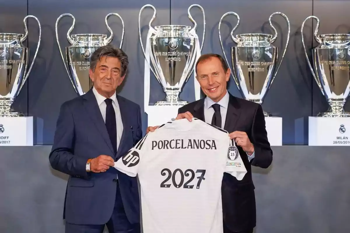 Two men in suits pose smiling, holding a white jersey with the name Porcelanosa and the number 2027 in front of several large football trophies. Two men in suits pose smiling, holding a white jersey with the name Porcelanosa and the number 2027 in front of several large football trophies.