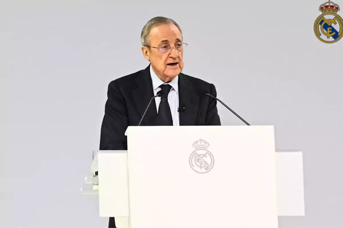 Older man in a dark suit speaking at a white podium with the Real Madrid crest in the background