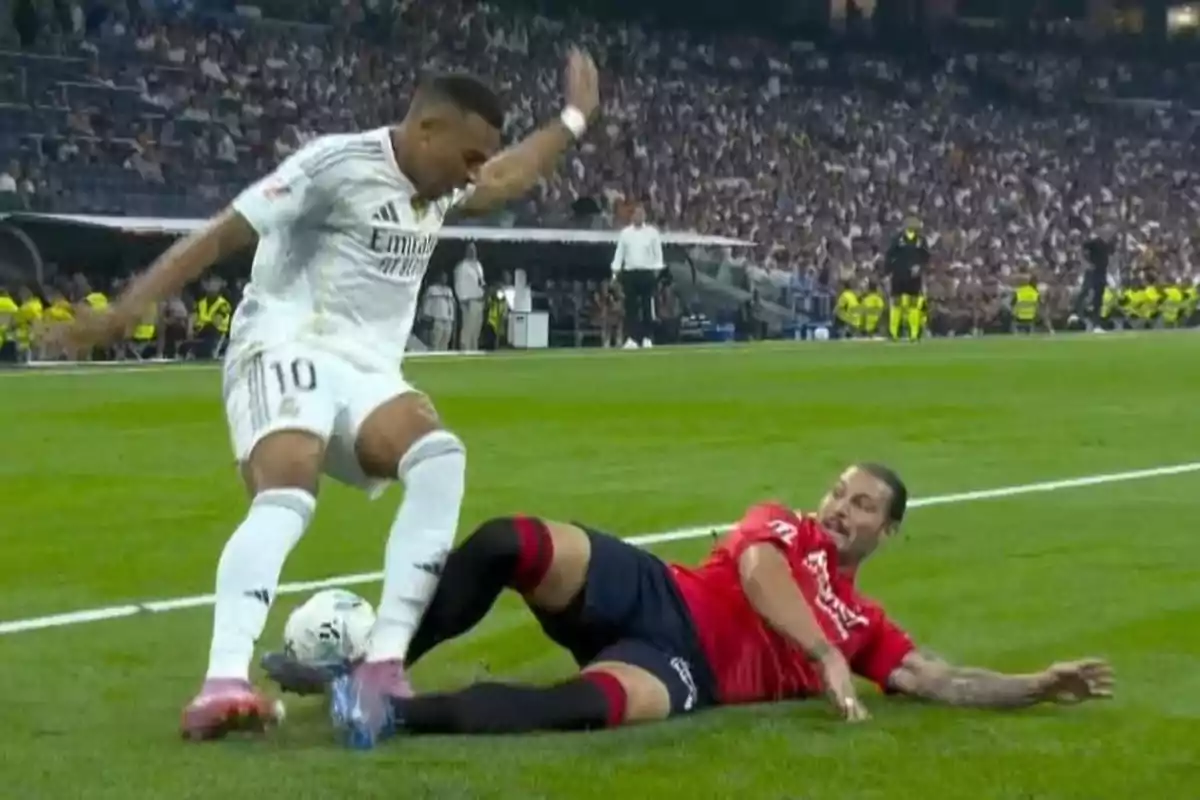 A player in a white uniform tries to dodge a slide tackle from an opponent dressed in red during a match in a stadium full of spectators. A player in a white uniform tries to dodge a slide tackle from an opponent dressed in red during a match in a stadium full of spectators.
