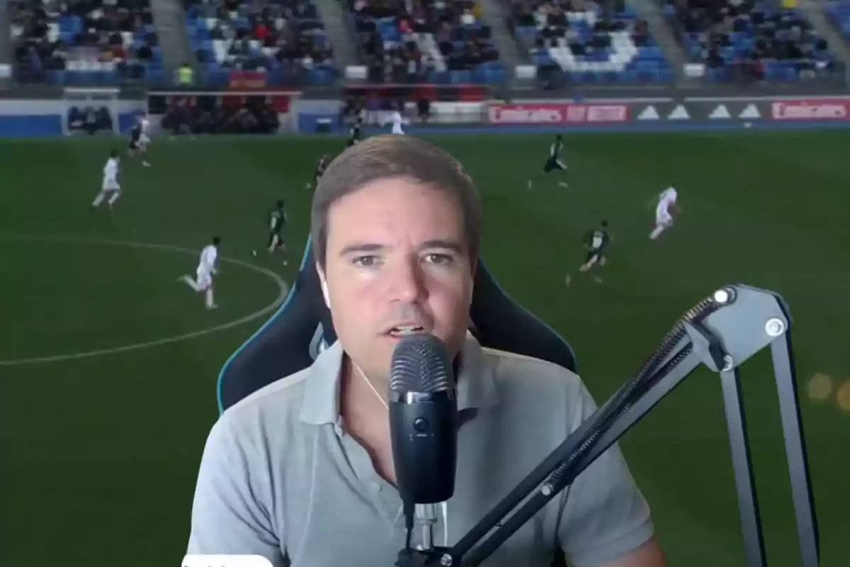 Hombre hablando frente a un micrófono con un partido de fútbol de fondo y público en las gradas