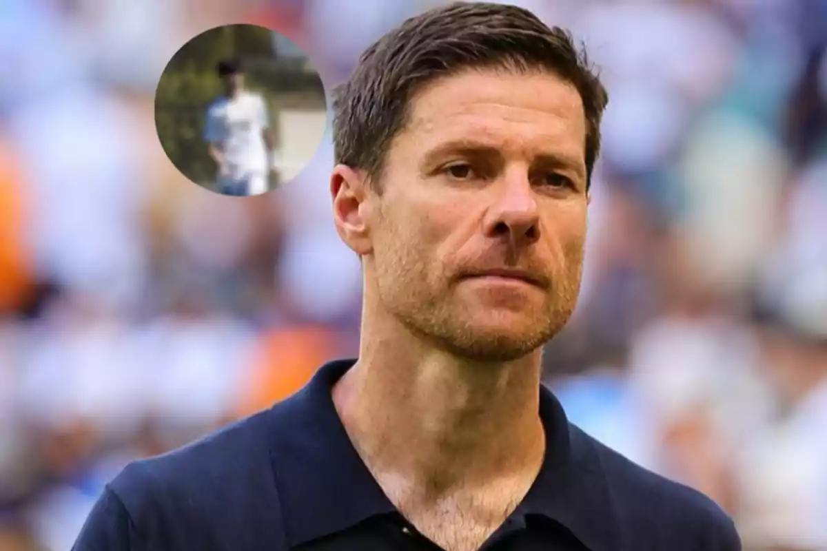 Hombre de cabello corto y barba ligera con expresión seria, viste una camiseta tipo polo azul oscuro y al fondo se observa un estadio desenfocado, en la esquina superior izquierda hay un círculo con la imagen borrosa de una persona vestida de blanco.