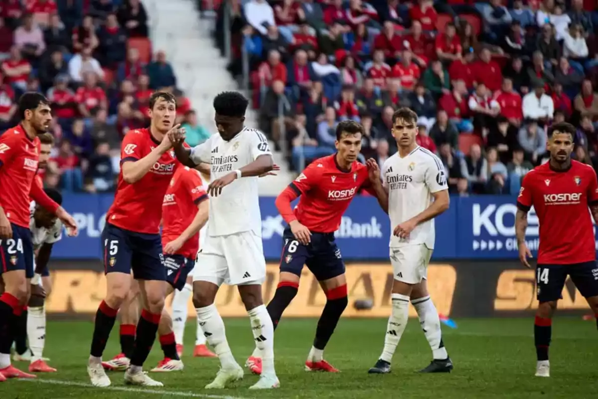 Players from two soccer teams, one wearing white uniforms and the other wearing red uniforms, get ready for a play in the box during a match with spectators in the stands.