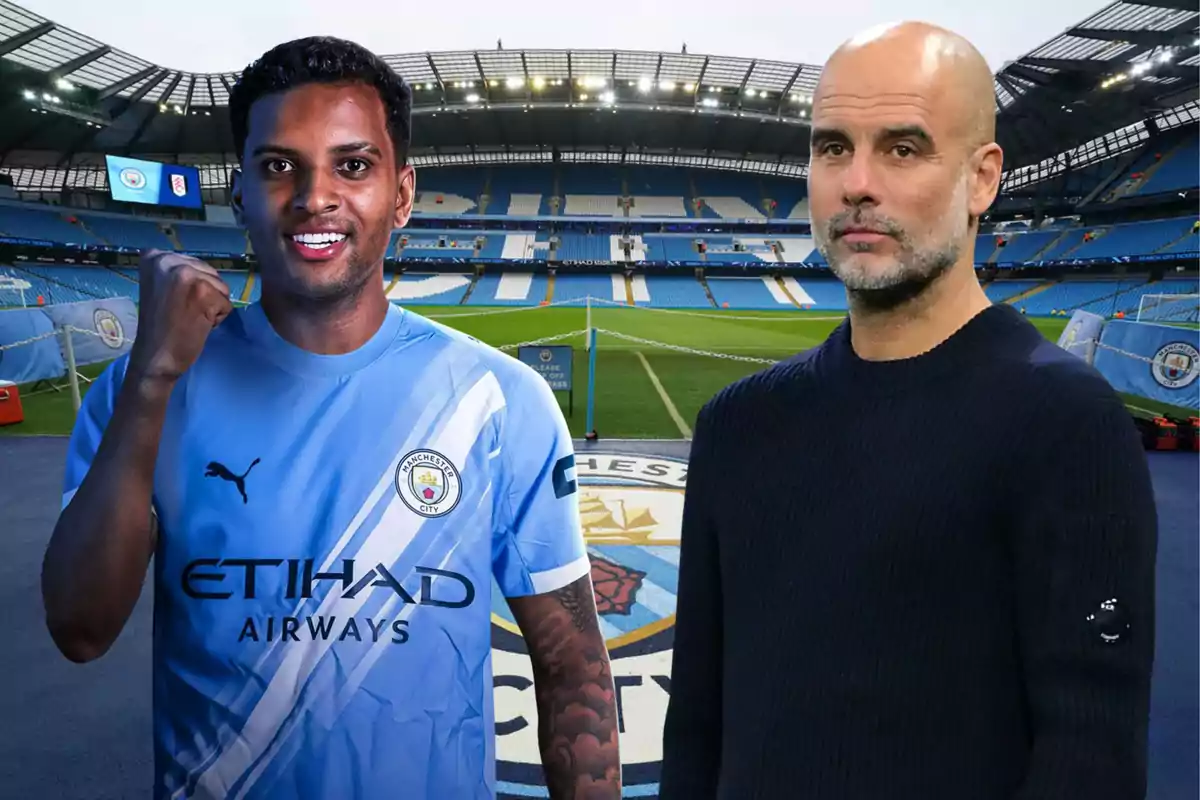 A Manchester City player smiles and raises his fist next to a bald man with a beard at the team's stadium, both posing on the field.